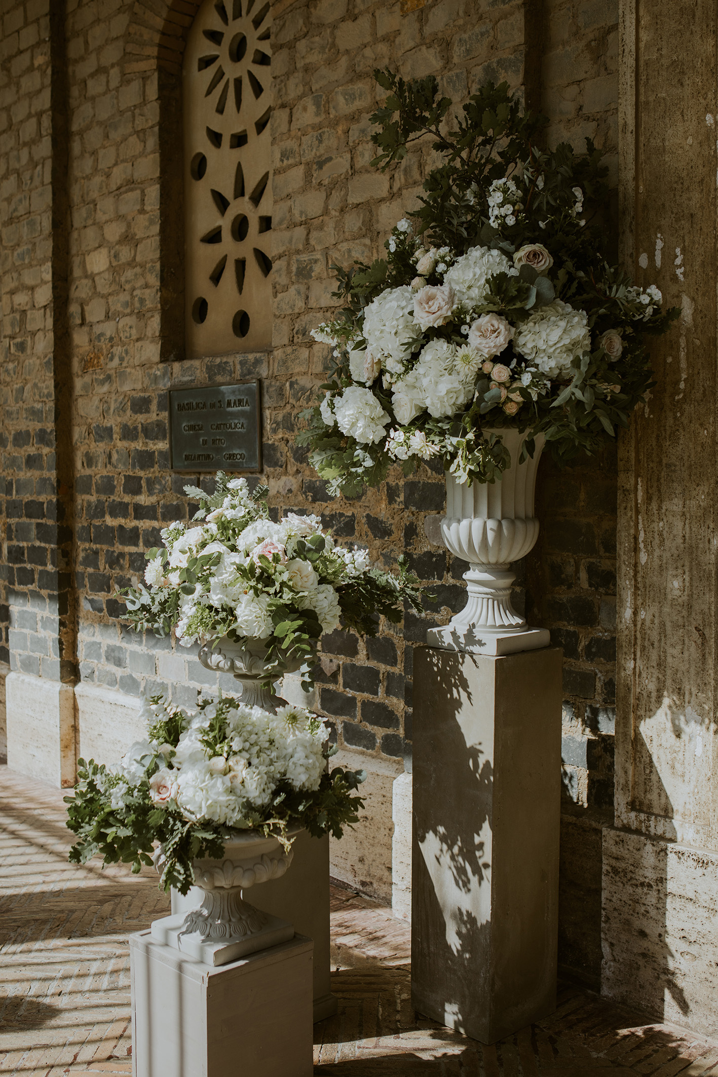 Addobbo floreale per ingresso chiesa matrimonio, con urne bianche e composizioni in stile botanico, verde di stagione e fiori bianchi
