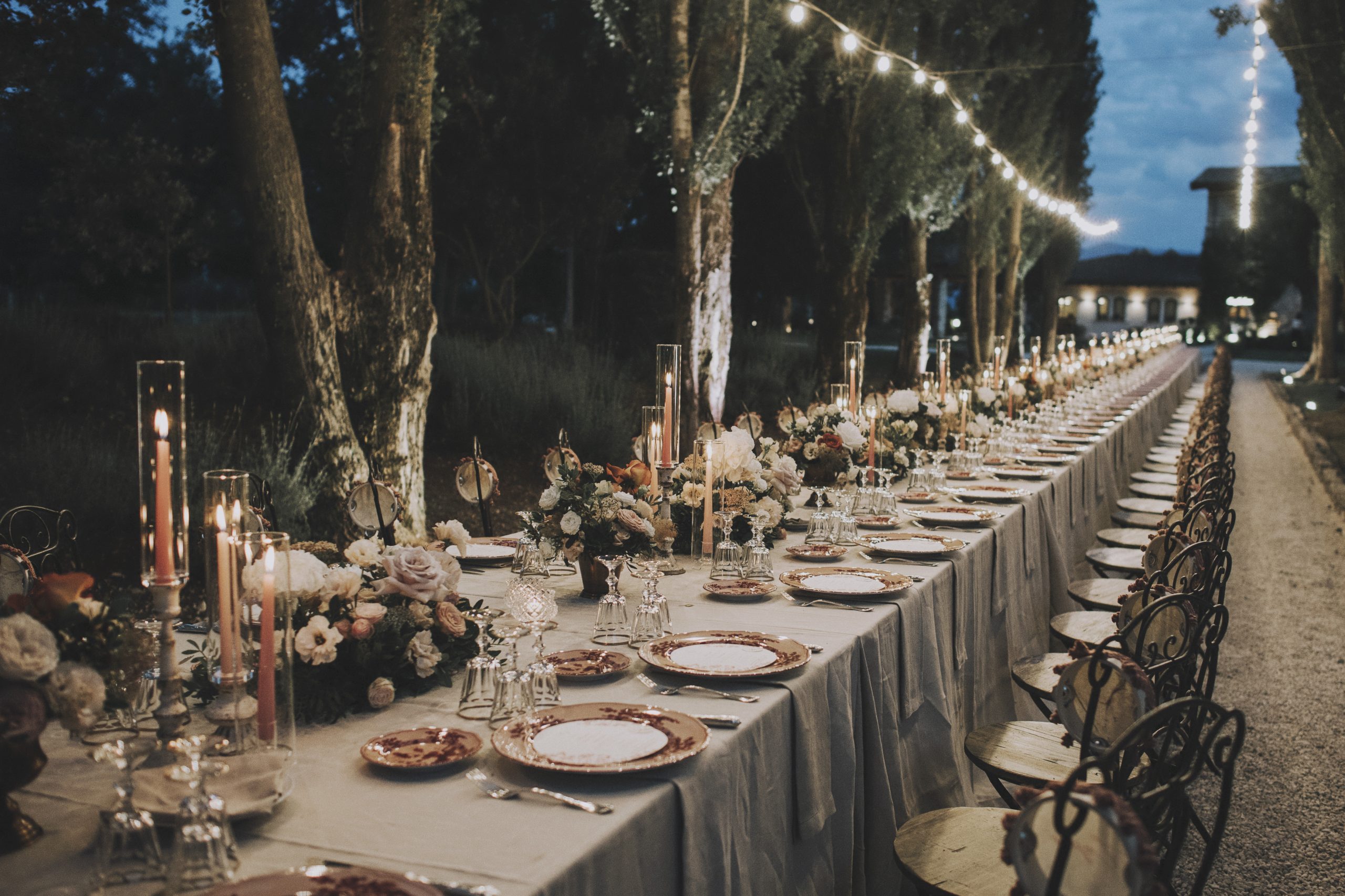 Decorazione per tavolo imperiale all'aperto con piccole composizioni di fiori nei toni del terracotta, rosa e bianco affiancate da candele alte rosa