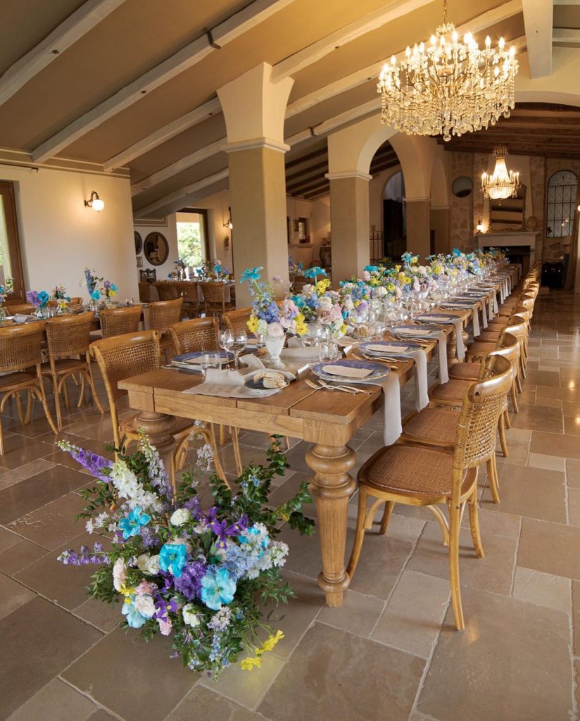 Tavolo imperiale in legno con centrotavola composto da piccole composizioni di fiori colorati in vasetti di cemento e decorazione floreale bassa ai piedi del tavolo